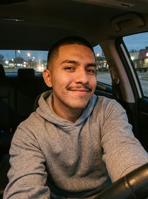 Front-camera selfie of a young man in a grey hoodie in a parked car