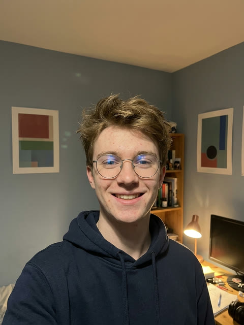 Bedroom selfie of a smiling young man with curly hair, round glasses and a navy hoodie