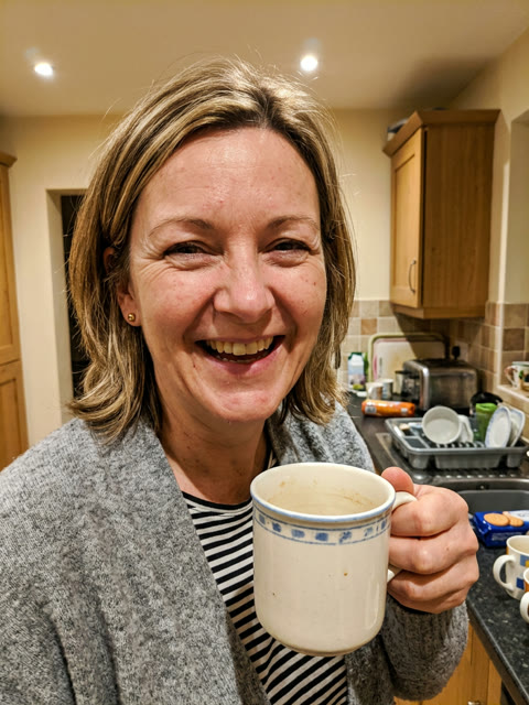 Kitchen selfie of a laughing woman with blonde hair in a grey cardigan holding a mug