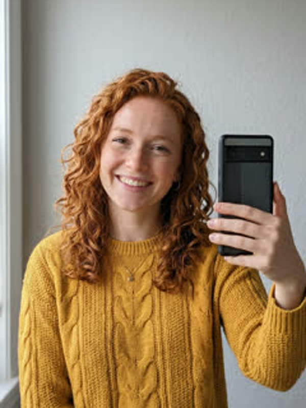 Mirror selfie of a smiling woman with curly red hair in a mustard cable-knit sweater