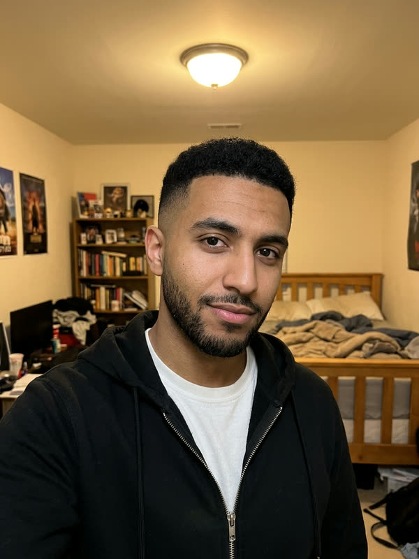 Bedroom selfie of a young man with a trimmed beard and a skin fade in a black zip hoodie over a white T-shirt